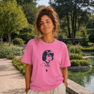 Op de afbeelding: Een felroze T-shirt met een zwart-wit grafisch portret. Het shirt met korte mouwen heeft een levendige roze kleur. De afbeelding is een gestileerd portret van een persoon. Het shirt wordt door een persoon in een buitenomgeving gedragen.