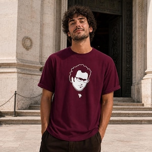 Op de afbeelding: Bordeauxrood T-shirt met een witte afbeelding van een persoon met een bril. Het T-shirt heeft korte mouwen en een ronde hals. De persoon op de foto draagt het T-shirt.