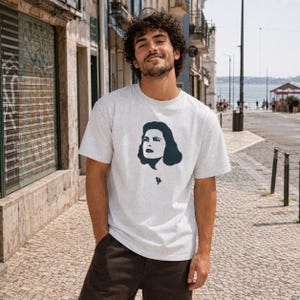 Op de afbeelding: Lichtgrijs t-shirt met een donkerblauwe afbeelding van een vrouwengezicht. Het t-shirt is gemaakt van een zacht materiaal en heeft een klassieke ronde hals. Het model draagt een bruine broek. De foto is buiten op een zonnige dag gemaakt.