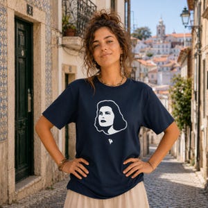Op de afbeelding: Marineblauw T-shirt met een witte afbeelding van een vrouwengezicht. Het T-shirt heeft korte mouwen en een ronde hals. De persoon op de foto draagt het T-shirt en staat buiten op een geplaveide straat met gebouwen op de achtergrond.