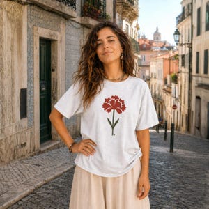 May include: A light gray t-shirt featuring a large, stylized red carnation flower with a green stem. The t-shirt is a crew neck style. The flower design is centered on the front of the shirt. The shirt is made of a soft material.