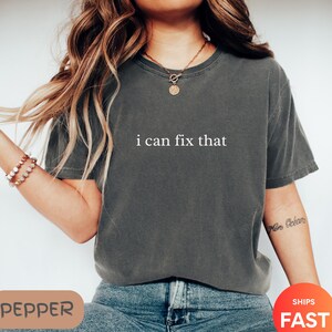 May include: Dark gray t-shirt with the phrase "i can fix that" printed in white. The shirt is a crew neck and has short sleeves. The model is wearing a necklace and bracelet.