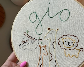 May include: Embroidered hoop art with the name "gio" in green thread. The hoop features a sloth hanging from a branch, two giraffes, and a lion. The background is a cream-colored fabric.