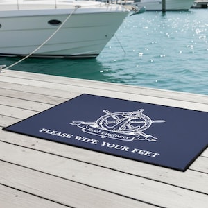 Puede incluir: Una alfombra rectangular azul marino con un diseño náutico blanco que presenta un timón, un ancla y una pancarta que dice "Reel Engineer". El texto "PLEASE WIPE YOUR FEET" está debajo. La alfombra está sobre un muelle de madera.
