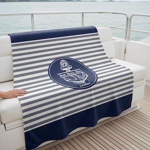 May include: A navy and white striped blanket with an anchor emblem. The blanket has a navy blue border and an oval design with an anchor graphic and the text "Boat Name Here" and "Location".