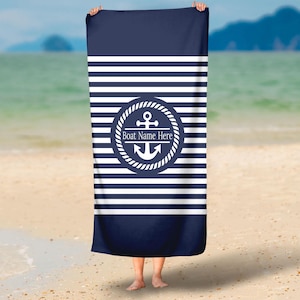 May include: A rectangular beach towel featuring a navy blue and white horizontal striped pattern. In the center, a circular emblem displays a white anchor surrounded by a rope border, with the text Boat Name Here printed in white.