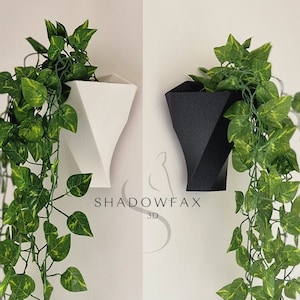 Puede incluir: Dos macetas de pared geométricas, una blanca y otra negra, con plantas verdes colgantes. Las macetas están hechas de un material impreso en 3D y tienen un diseño único y moderno. El texto "SHADOWFAX 3D" es visible debajo de las macetas.
