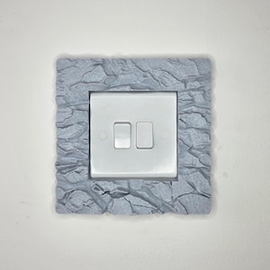 May include: A white double light switch mounted within a square, grey, textured frame that mimics the appearance of jagged, cracked rock or stone. The switch plate features two vertical toggles and a smooth, clean finish on the central component.