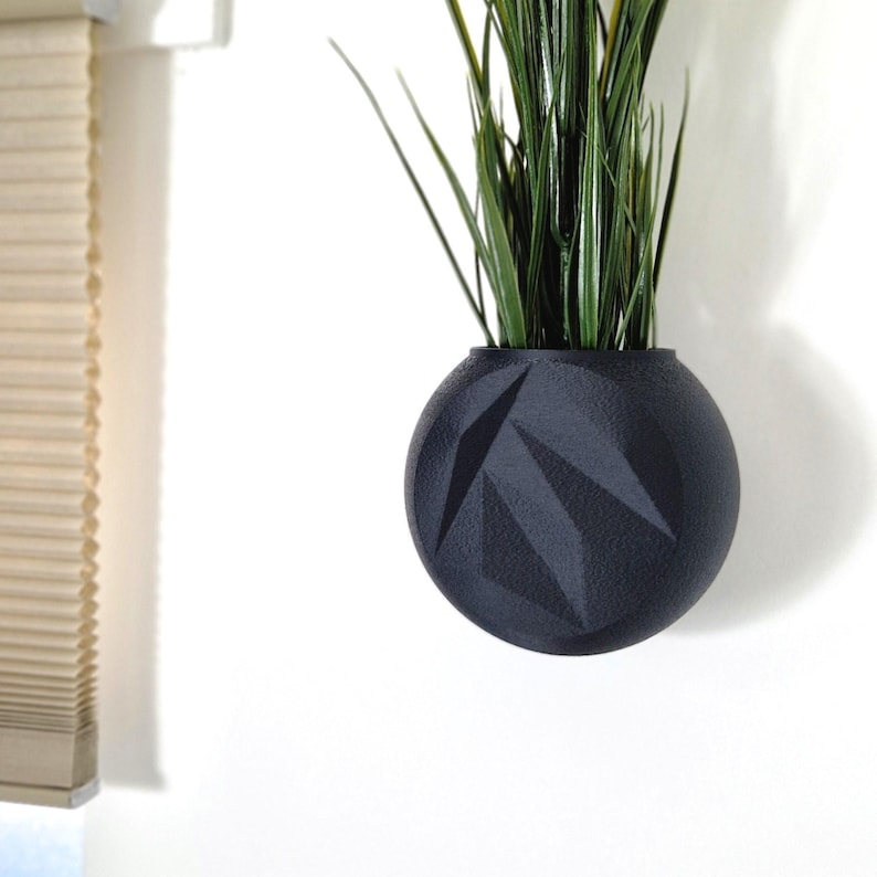 May include: A black geometric planter with a green plant inside. The planter is hanging on a white wall.