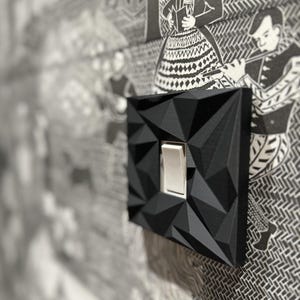 May include: A black light switch cover plate featuring a geometric, faceted, three-dimensional surface texture. The cover plate is square-shaped with a central rectangular cutout containing a standard white toggle light switch mechanism.