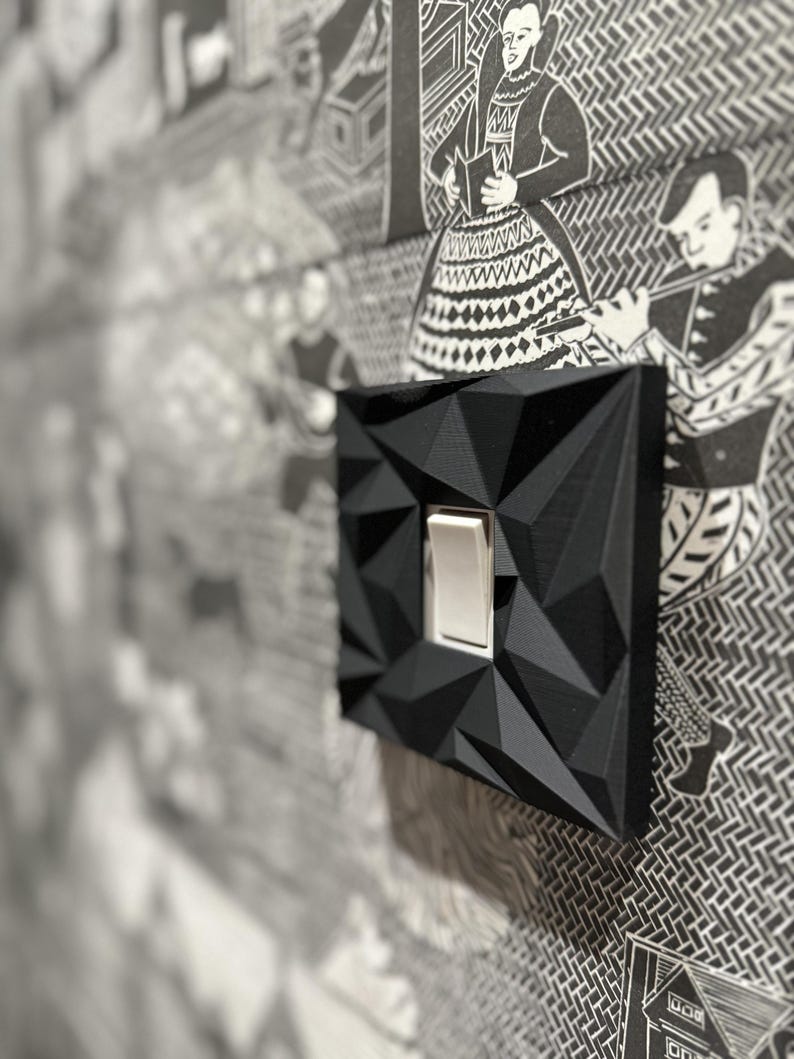 May include: A black light switch cover plate featuring a geometric, faceted, three-dimensional surface texture. The cover plate is square-shaped with a central rectangular cutout containing a standard white toggle light switch mechanism.