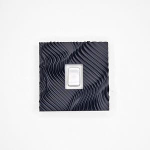 May include: A square light switch cover plate featuring a dark navy blue finish with a textured, wavy, topographical line pattern across the surface. A single, rectangular, white rocker-style light switch is centered within the plate.