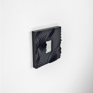 May include: A square black light switch cover plate featuring a deep, wavy, three-dimensional texture across its surface. The cover plate is mounted on a wall and surrounds a standard white rectangular rocker light switch in the center.