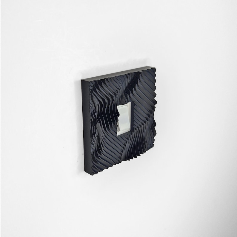 May include: A square black light switch cover plate featuring a deep, wavy, three-dimensional texture across its surface. The cover plate is mounted on a wall and surrounds a standard white rectangular rocker light switch in the center.