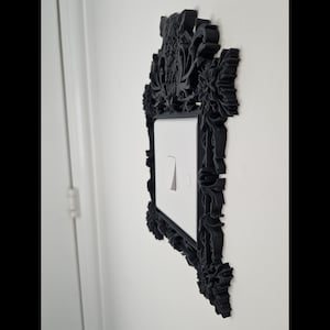 May include: Black decorative light switch cover with ornate floral and scrollwork detailing. The cover surrounds a white light switch with a silver toggle. The design is mounted on a white wall.