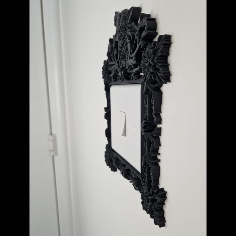 May include: Black decorative light switch cover with ornate floral and scrollwork detailing. The cover surrounds a white light switch with a silver toggle. The design is mounted on a white wall.