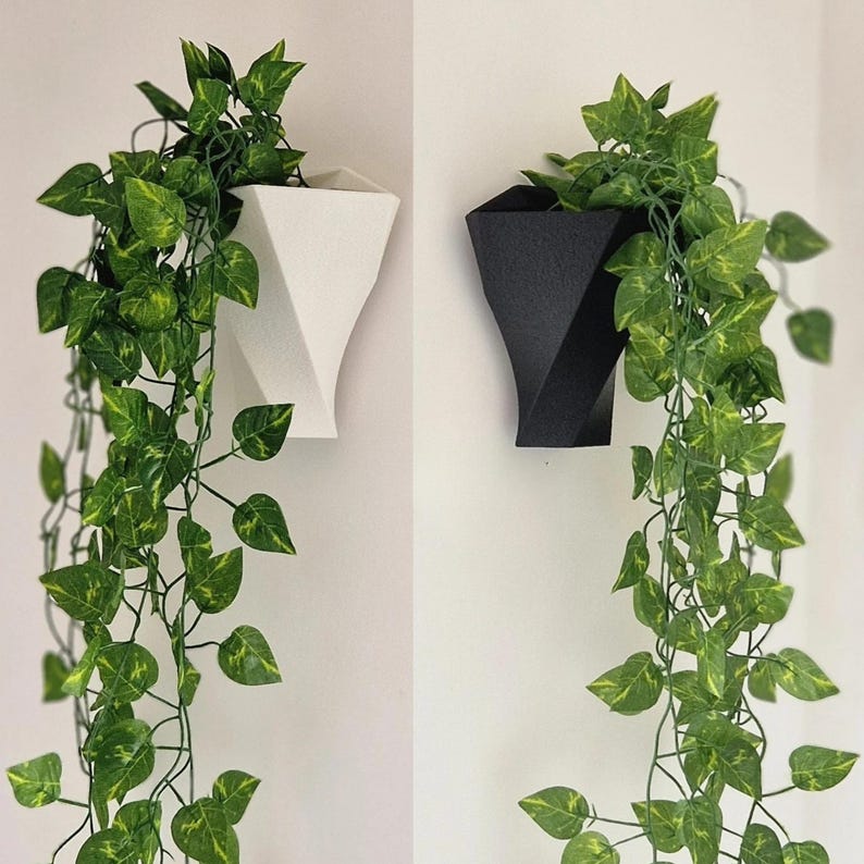 May include: Two wall-mounted planters, one white and one black, each featuring a geometric, faceted design. Both planters contain trailing artificial green ivy vines with variegated leaves that drape downwards from the open top openings.
