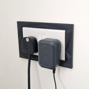 Może przedstawiać: Ciemnoszara, geometryczna ramka na gniazdko elektryczne otaczająca białe podw&oacute;jne gniazdo. Czarna wtyczka z trzema bolcami jest włożona do lewego gniazda, a ciemnoszary zasilacz marki Shark jest podłączony do prawego gniazda z przewodami.