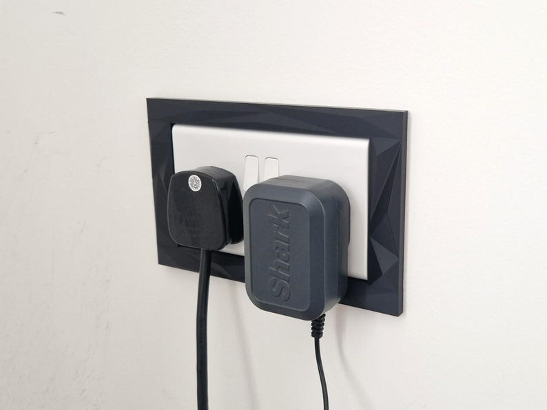 Może przedstawiać: Ciemnoszara, geometryczna ramka na gniazdko elektryczne otaczająca białe podw&oacute;jne gniazdo. Czarna wtyczka z trzema bolcami jest włożona do lewego gniazda, a ciemnoszary zasilacz marki Shark jest podłączony do prawego gniazda z przewodami.