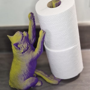 May include: A purple and yellow cat figurine holding a roll of white toilet paper. A small bird figurine is perched on top of the toilet paper roll.