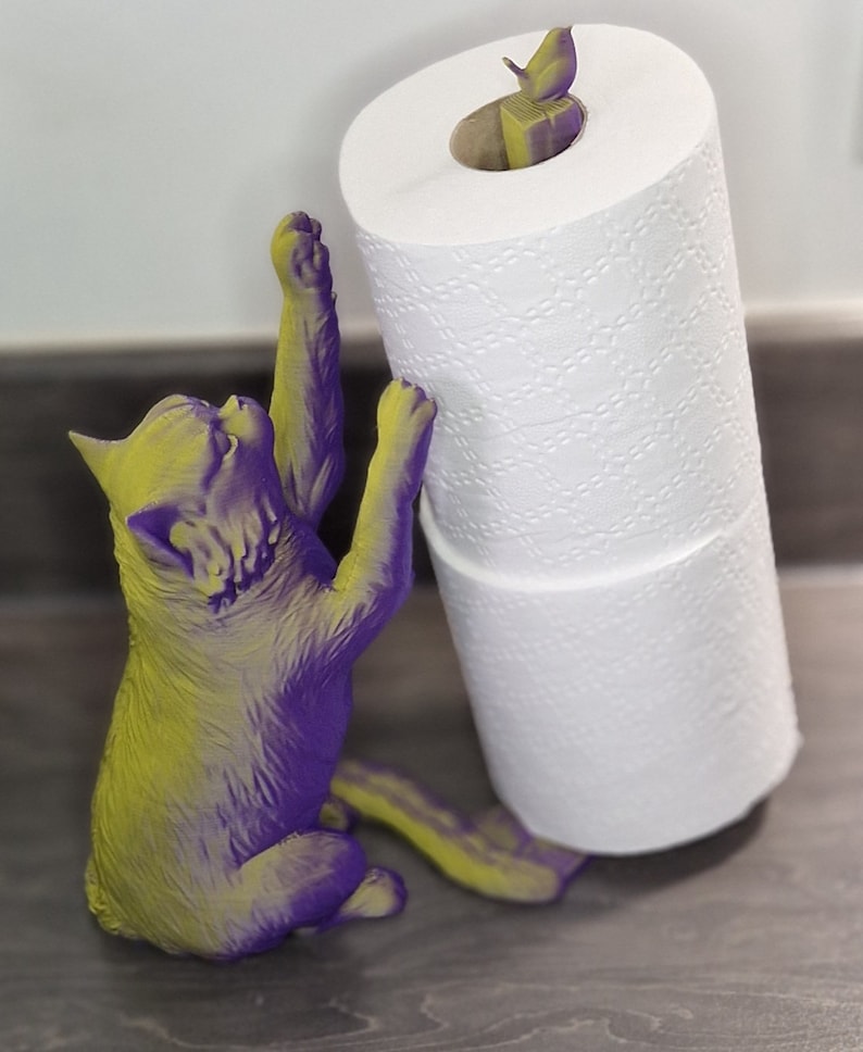 May include: A purple and yellow cat figurine holding a roll of white toilet paper. A small bird figurine is perched on top of the toilet paper roll.