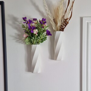 May include: Two white ceramic vases with a textured, twisted design. One vase holds a bouquet of purple and white flowers, while the other holds dried beige grasses.