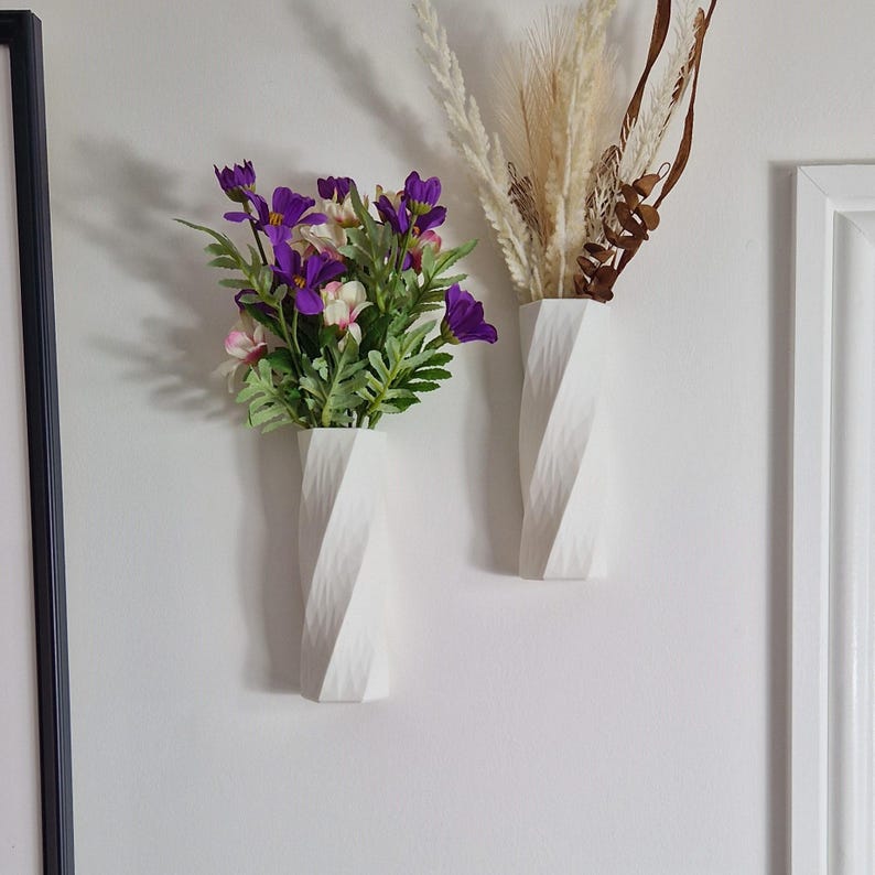 May include: Two white ceramic vases with a textured, twisted design. One vase holds a bouquet of purple and white flowers, while the other holds dried beige grasses.