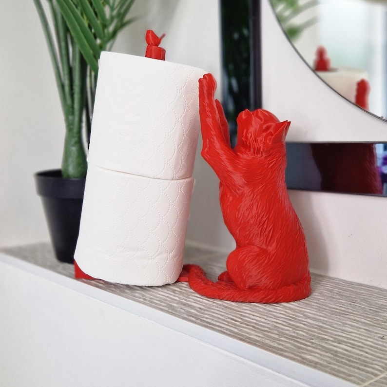 May include: Red cat-shaped toilet paper holder with a roll of white toilet paper. The cat is holding the toilet paper roll with its paws.