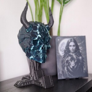 May include: A teal blue and black 3D printed skull with horns on a black stand. The skull has a detailed, textured surface.