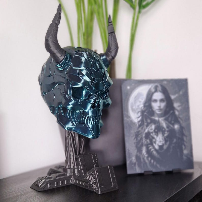 May include: A teal blue and black 3D printed skull with horns on a black stand. The skull has a detailed, textured surface.