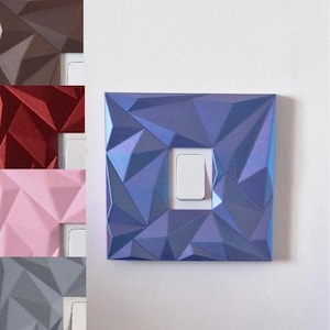 May include: A square light switch cover plate featuring a geometric, faceted, low-poly surface texture in a metallic blue finish. The central rectangular white toggle switch is surrounded by the angular, multi-planar blue frame design.