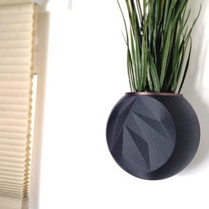 May include: A black geometric planter with a copper rim, hanging on a white wall. The planter is filled with green plants.