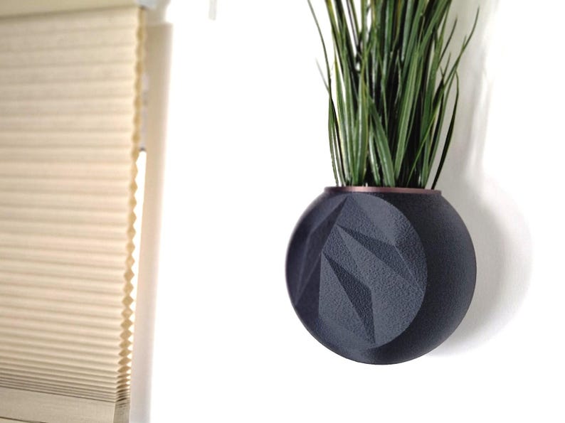 May include: A black geometric planter with a copper rim, hanging on a white wall. The planter is filled with green plants.