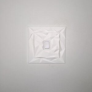 May include: A white light switch with a geometric, faceted design. The switch plate is square with a central rectangular switch. The design features a series of triangular shapes, creating a textured appearance. The switch is in the off position.
