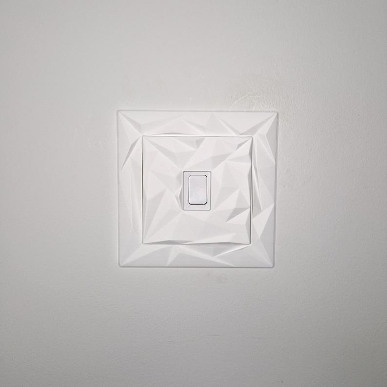 May include: A white light switch with a geometric, faceted design. The switch plate is square with a central rectangular switch. The design features a series of triangular shapes, creating a textured appearance. The switch is in the off position.
