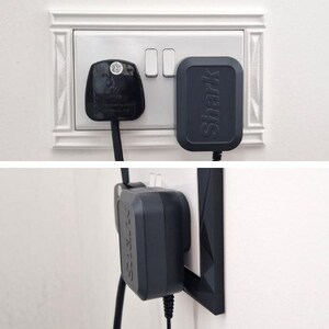 May include: A black Shark brand power adapter plugged into a white wall outlet alongside a black three-prong plug. The adapter features a rectangular shape with rounded corners and the word Shark embossed vertically on its front surface.