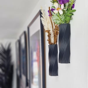 May include: Two black, twisted, ceramic wall vases with dried flowers and a bouquet of purple and white flowers.