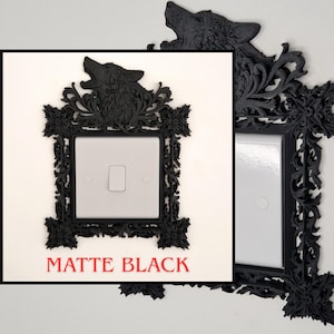 May include: Matte black light switch cover with a decorative frame. The frame features a howling wolf at the top and ornate floral detailing. The text "MATTE BLACK" is printed in red at the bottom.