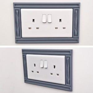 May include: A white double electrical wall outlet with two switches, framed by a decorative gray rectangular border featuring a textured, molded edge design. The outlet is shown from both a direct front view and an angled perspective.