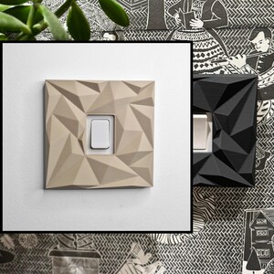May include: A square light switch cover plate featuring a geometric, faceted, low-poly surface design. The plate is available in a matte beige finish with a central white toggle switch and a matte black finish with a white toggle switch.