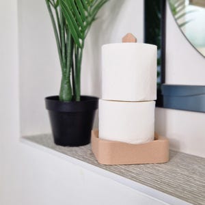 Puede incluir: Dos rollos de papel higi&eacute;nico blanco apilados en un soporte de madera marr&oacute;n claro con un peque&ntilde;o pomo de madera en la parte superior. El soporte est&aacute; sobre una encimera blanca.