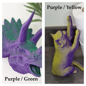 May include: Two 3D printed figurines, one of a purple and green dinosaur and one of a purple and yellow cat. The dinosaur is a Triceratops with a large horn on its nose and two smaller horns above its eyes. The cat is standing on its hind legs with its front paws raised. The text "Purple / Green" is below the dinosaur and "Purple / Yellow" is below the cat.