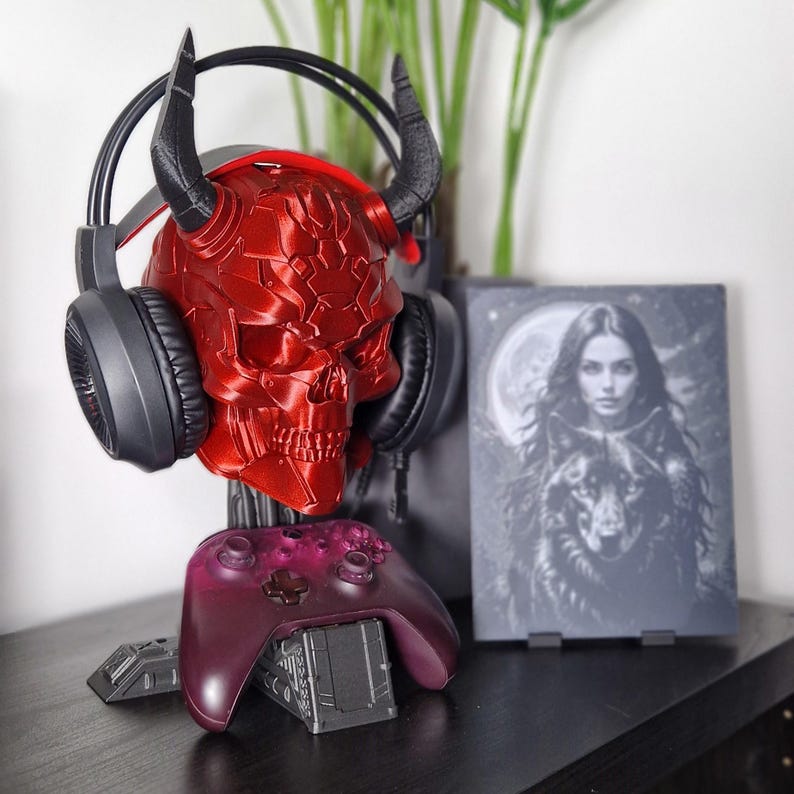 May include: A red skull-shaped headset stand with a black headset and a purple video game controller. The stand is on a black surface with a framed print of a woman and a wolf.