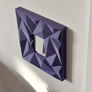 May include: A square light switch cover plate featuring a dark purple, three-dimensional geometric faceted surface. The cover plate surrounds a single, centered white rocker-style light switch, creating a textured, angular appearance on the wall.