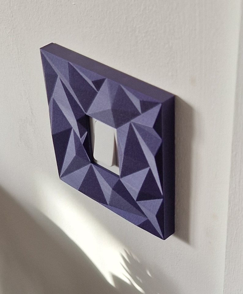 May include: A square light switch cover plate featuring a dark purple, three-dimensional geometric faceted surface. The cover plate surrounds a single, centered white rocker-style light switch, creating a textured, angular appearance on the wall.