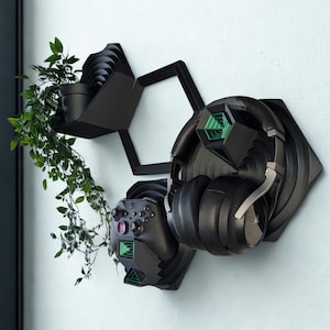 Könnte beinhalten: Ein schwarzer, an der Wand montierter Gaming-Organizer mit einem Regal, das eine Topfpflanze und ein sechseckiges Design enthält. Er verfügt über eine Halterung für einen Gaming-Controller und ein Headset. Das Headset ist schwarz mit silbernen Akzenten.