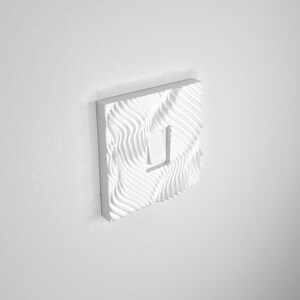 May include: A square white light switch cover plate featuring a textured, wavy, rippled surface pattern. The cover plate is mounted on a wall and surrounds a single, centered, rectangular white toggle switch mechanism in the middle.