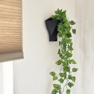 May include: A matte black geometric wall planter with a textured surface holding a trailing artificial vine. The plant features numerous small, pointed green leaves with light yellow-green variegation that cascade downward from the container.