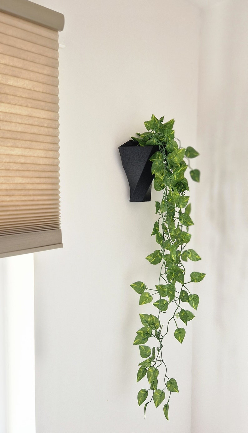 May include: A matte black geometric wall planter with a textured surface holding a trailing artificial vine. The plant features numerous small, pointed green leaves with light yellow-green variegation that cascade downward from the container.