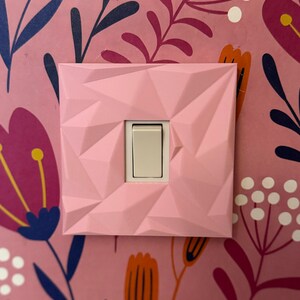 May include: A square light switch cover with a faceted, geometric pink surface texture. The cover features a central rectangular opening that reveals a standard off-white toggle switch mechanism, mounted against a pink floral wallpaper background.